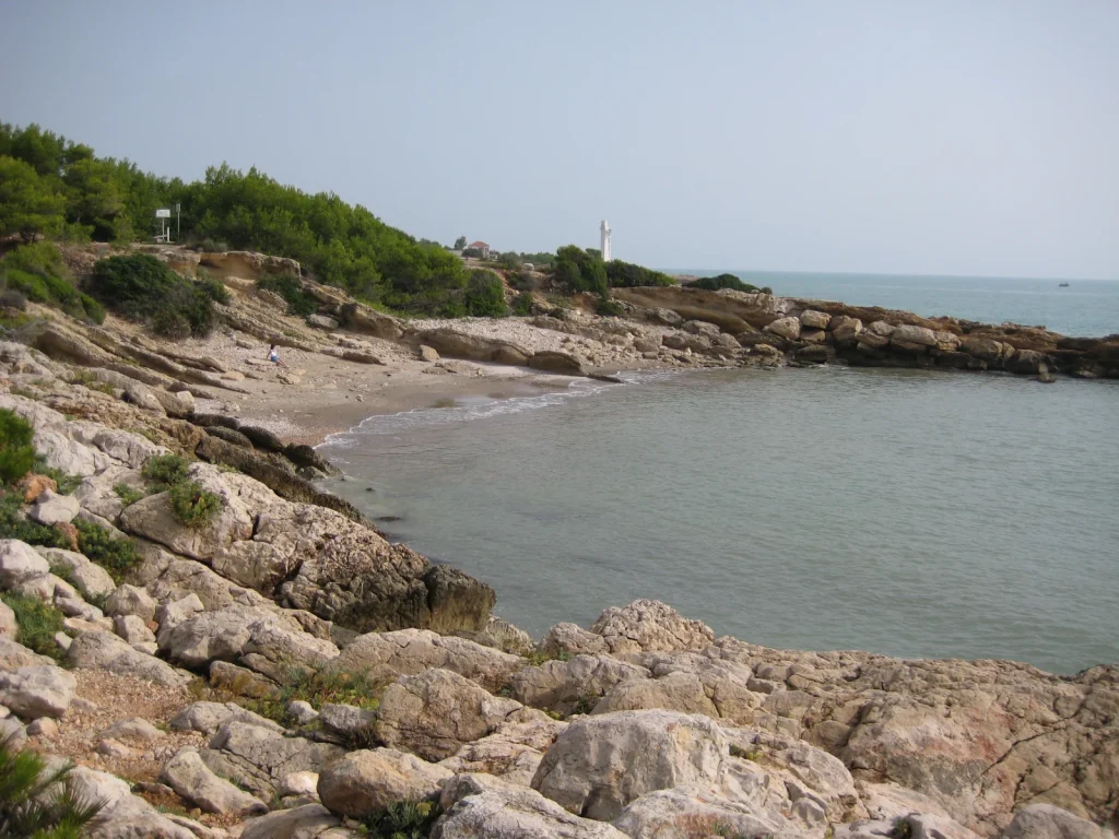 Cala Blanca en Alcossebre, rodeada de rocas y pinos, con el faro de Irta al fondo